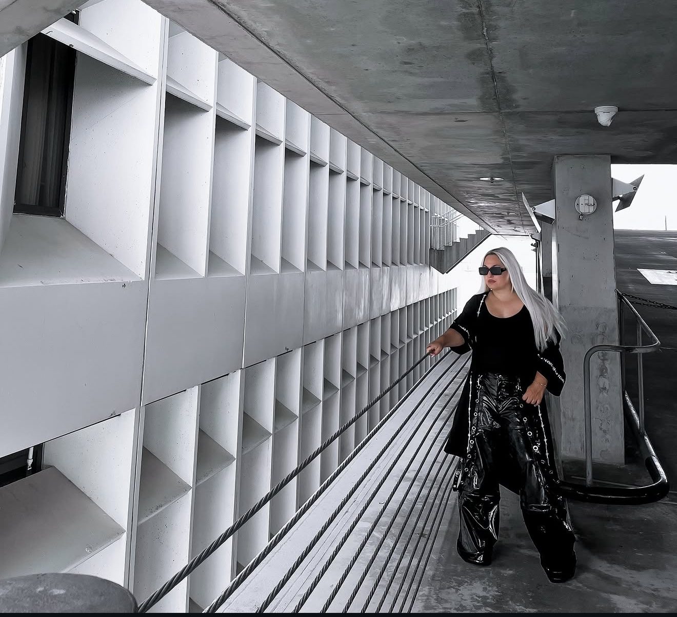 TENAJ photographed in a concrete walkway, in black patent trousers and sunglasses.